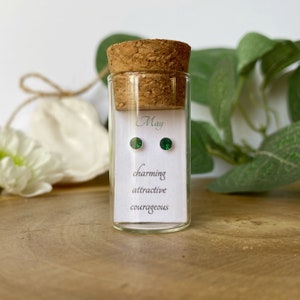 Könnte beinhalten: Ein klares Glasfläschchen mit Korken enthält ein Paar kleiner, runder, smaragdgrüner Ohrstecker. Eine weiße Karte im Inneren des Fläschchens trägt die Aufschrift "May", "charming", "attractive" und "courageous". Das Fläschchen steht auf einer Holzoberfläche.
