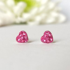 Könnte beinhalten: Ein Paar herzförmige Ohrstecker. Jeder Ohrring hat eine Ansammlung leuchtend rosa Kristalle, die in einem silberfarbenen Metall gefasst sind. Die Ohrringe werden vor einem weichen, verschwommenen Hintergrund präsentiert.