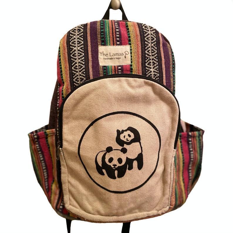 May include: A multi-colored striped backpack with a large white canvas front pocket featuring a black and white panda bear design. The text "The Lamas Handmade in Nepal" is printed on a small tag attached to the backpack.