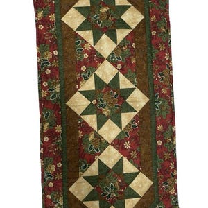 May include: A quilted table runner featuring a pointed end design with a red fabric border patterned with green holly leaves and small flowers. The center panel displays three star-shaped quilt blocks in cream and dark green fabric.