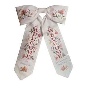 Puede incluir: Lazo de bienvenida de punto de cruz color crema con bordado floral. La palabra "WELCOME" está bordada en rojo, con el apellido "The Rosenfelds" y la dirección "24 Sycamore Waltham" debajo.