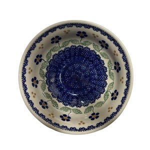 May include: A round ceramic bowl featuring a central dark blue field with white stippling, surrounded by a ring of light green leaves and small dark blue flowers with yellow centres. The rim has a decorative dark blue scalloped pattern.