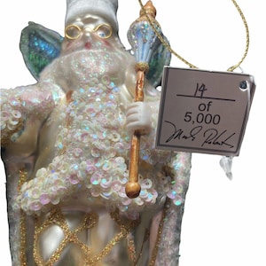 Op de afbeelding: Een decoratieve kerstversiering die een feeachtig figuur afbeeldt met een witte hoed, een goudkleurige bril en een staf. Het figuur is versierd met pailletten en glitter, en heeft iriserende vleugels. Een label leest "14 of 5,000".