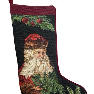 May include: A needlepoint Christmas stocking featuring a black background with a central portrait of Santa Claus wearing a red hat and coat. The design includes green pine boughs, red berries, and a poinsettia motif at the toe with a red fabric trim.