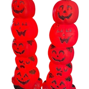 Può includere: Due decorazioni di zucche rosse impilate di Halloween con facce intagliate. Ogni pila di zucche ha cinque zucche, con uno stelo in cima e una base nera. Le zucche hanno facce sorridenti intagliate e sono illuminate dall'interno.