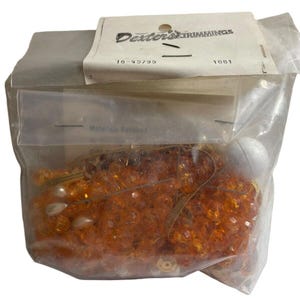 May include: A clear plastic bag filled with orange and gold beads, gold ribbon, and a white pearl. The bag has a label that reads "Dexter's Trimmings" with the numbers "16-45795" and "1081".