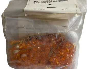 Könnte beinhalten: Eine durchsichtige Plastiktüte gefüllt mit orangefarbenen und goldenen Perlen, goldfarbenem Band und einer weißen Perle. Die Tüte trägt ein Etikett mit der Aufschrift "Dexter's Trimmings" und den Nummern "16-45795" und "1081".
