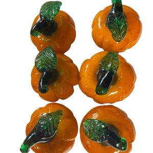 Può includere: Sei zucche di vetro arancione con steli e foglie verdi su sfondo bianco. Queste zucche lucide sono probabilmente oggetti decorativi per l'autunno o Halloween.