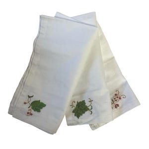 Pode incluir: Um conjunto de três guardanapos de tecido branco dobrados juntos, cada um com um design bordado distinto de folhas verdes e pequenas bagas vermelhas no canto, exibindo costuras detalhadas e uma textura de tecido limpa e nítida.