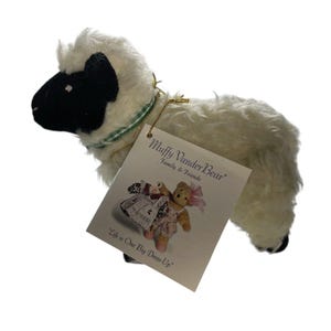 May include: A plush sheep toy with a black face and feet, and a white body. It has a green and gold collar and a tag that reads "Muffy VanderBear Family & Friends" and "Life is One Big Dress-Up".