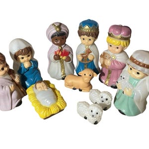 May include: A ceramic nativity set featuring figures of the baby Jesus, Mary, Joseph, the three wise men, and several sheep. The figures are painted in pastel colors with gold accents. The baby Jesus is in a yellow manger.