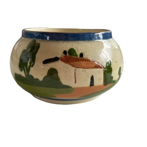 May include: A round, hand-painted ceramic bowl with a landscape design. The bowl features a cream-colored base with a blue rim and a scene of a house, trees, and hills in green, brown, and orange.