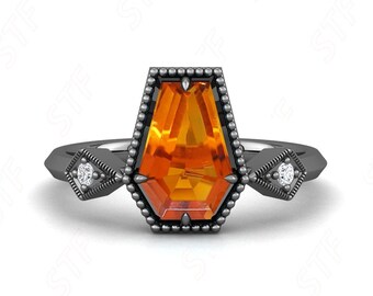 May include: A black ring featuring an orange gemstone in a coffin shape. The ring has a beaded border and small diamonds on either side. The ring is set against a white background.