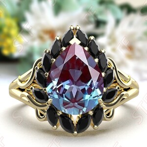 May include: A pear-shaped ring with a large, multi-colored gemstone surrounded by smaller black gemstones. The ring is made of gold and has a delicate, ornate design.