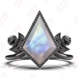 Puede incluir: Un anillo negro con una gran piedra preciosa iridiscente y facetada engastada en un metal negro. La piedra preciosa está rodeada de piedras preciosas negras más pequeñas. El anillo está sobre un fondo blanco.