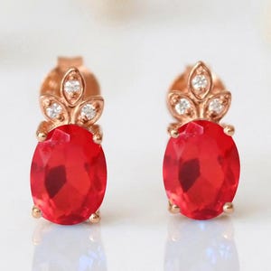 May include: A pair of rose gold stud earrings featuring a large oval-cut red gemstone as the centerpiece. Above each red stone, three small clear accent stones are arranged in a leaf-like cluster, secured by metal prongs on a rose gold base.