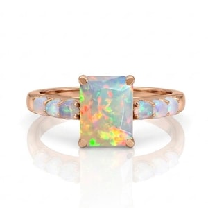 May include: A rose gold ring featuring a large rectangular emerald-cut opal as the center stone. The band is accented with three smaller round opal stones on each side of the central setting, showcasing iridescent flashes of green, orange, and yellow.