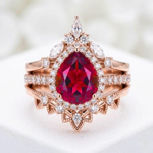 May include: A rose gold ring set featuring a large, pear-shaped, deep red gemstone surrounded by a halo of small, clear gemstones. Additional clear gemstones adorn the band and crown the ring, creating a luxurious and elegant design.