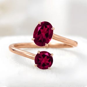 May include: A bypass ring crafted from rose-colored metal featuring two prong-set gemstones. One stone is oval-shaped and the other is round, both displaying a deep, saturated magenta-red hue with visible internal facets and light reflection.