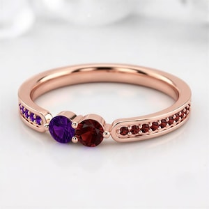 May include: A rose gold ring featuring two prominent round-cut stones, one purple amethyst and one deep red garnet, set side-by-side. The band is accented with a channel of smaller purple stones on one side and red stones on the other.