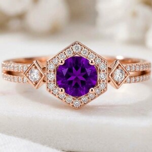 May include: A rose gold ring featuring a round purple amethyst center stone set within a hexagonal halo of small white diamonds. The band includes two diamond-accented square settings on either side and a split shank design with pavé diamonds.