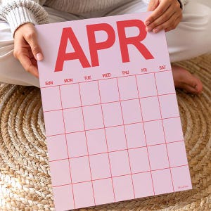 May include: A pink April calendar with red lettering and grid lines. The days of the week are listed across the top. The calendar is held by a person.