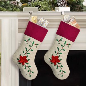 Puede incluir: Dos calcetines de Navidad beige con puños de fieltro rojo y flores de poinsettia y hojas de acebo bordadas. Los calcetines están colgados en una repisa de chimenea blanca.