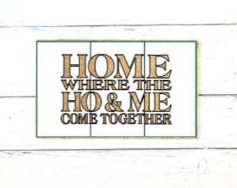 May include: A rectangular wall sign with a white background and a thin green border. The sign features the black text "HOME WHERE THE HO & ME COME TOGETHER" in a bold, serif font. The design is divided into three vertical panels by thin lines.