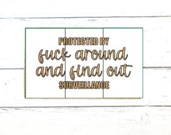 May include: A rectangular white sign with a dark green border featuring the text PROTECTED BY fuck around and find out SURVEILLANCE in gold lettering. The sign is divided into three vertical panels by thin black lines across the surface.