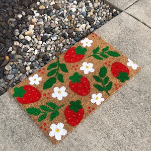 May include: A brown coir doormat with a white and green strawberry and flower design. The doormat is on a gray concrete surface.