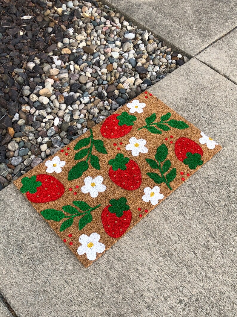 May include: A brown coir doormat with a white and green strawberry and flower design. The doormat is on a gray concrete surface.