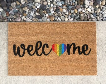 Può includere: Un tappetino per porta in fibra di cocco marrone con la parola "welcome" in caratteri neri e un cuore arcobaleno al centro.