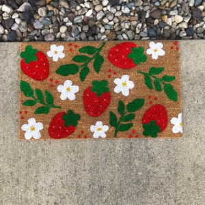 May include: A brown coir doormat with a strawberry and flower design. The strawberries are red with green leaves and the flowers are white with yellow centers.