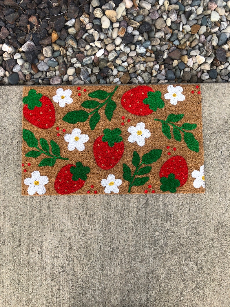 May include: A brown coir doormat with a strawberry and flower design. The strawberries are red with green leaves and the flowers are white with yellow centers.