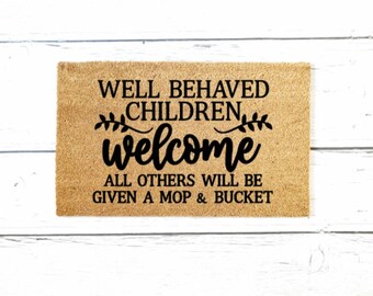 Può includere: Un tappetino per porta in fibra di cocco marrone con il testo "Well behaved children welcome. All others will be given a mop & bucket." in lettere nere.