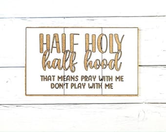 May include: A rectangular white wooden sign with three vertical panels. The text "HALF HOLY half hood" is printed in large, glittery gold letters, followed by the black text "THAT MEANS PRAY WITH ME DON'T PLAY WITH ME" centered below.