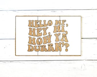 May include: A rectangular doormat with a natural tan fiber texture featuring the brown printed text: HELLO MF, HEY, HI, HOW YA DURRN? The lettering is arranged in a stacked, wavy, retro-inspired font style across the center of the mat.