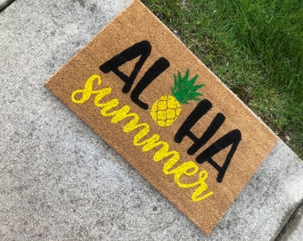 Può includere: Un tappetino per porta in fibra di cocco marrone con il testo "Aloha Summer" in lettere nere e gialle. Un ananas giallo con foglie verdi è centrato tra le parole.