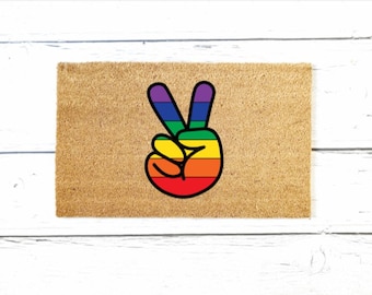 May include: A brown doormat with a rainbow peace sign. The peace sign is a hand with two fingers raised in the peace sign gesture.