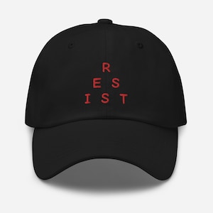Op de afbeelding: Zwarte baseballpet met rode geborduurde tekst die "RESIST" in een driehoekig patroon leest.