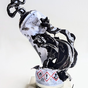 Puede incluir: Escultura de un bote de pintura en aerosol con pintura negra brillante que parece fluir. El bote es blanco con detalles en negro y gris, y la parte superior es blanca. La escultura se encuentra sobre una pequeña base cuadrada de hormigón.