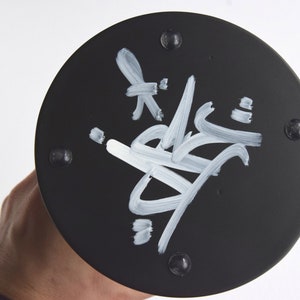 May include: Black round base with a white signature on it. The base has six small black dots around the edge.