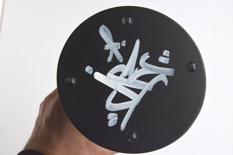 May include: Black round base with a white signature on it. The base has six small black dots around the edge.