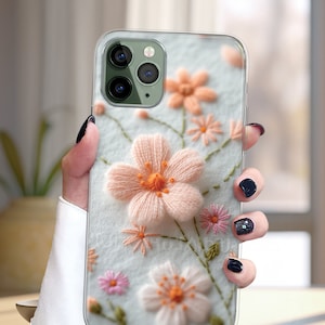 May include: A clear phone case with a floral embroidery design. The flowers are pink, white, and green. The case is designed for an iPhone.