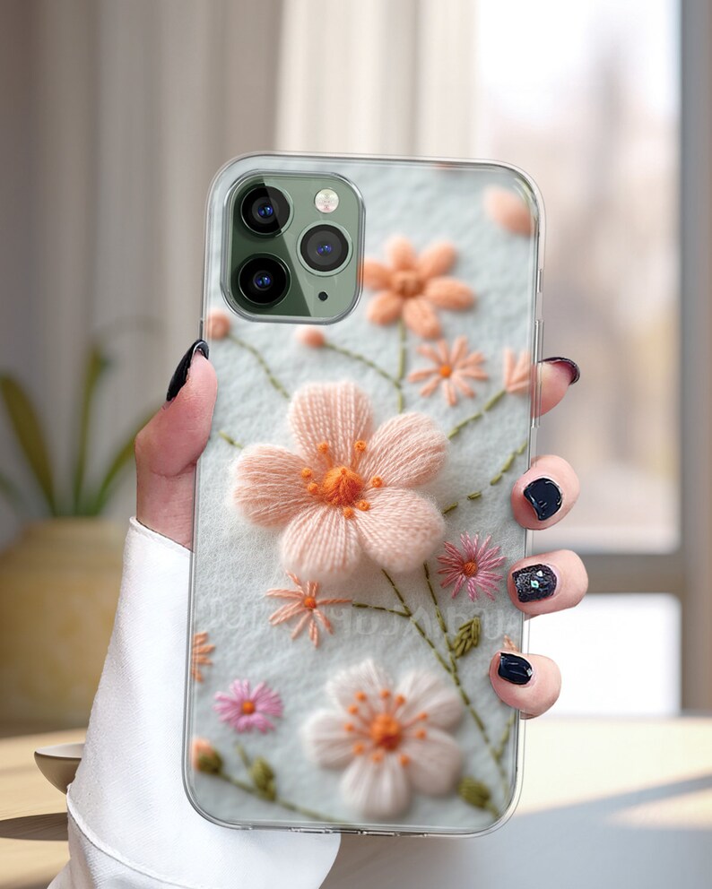 May include: A clear phone case with a floral embroidery design. The flowers are pink, white, and green. The case is designed for an iPhone.