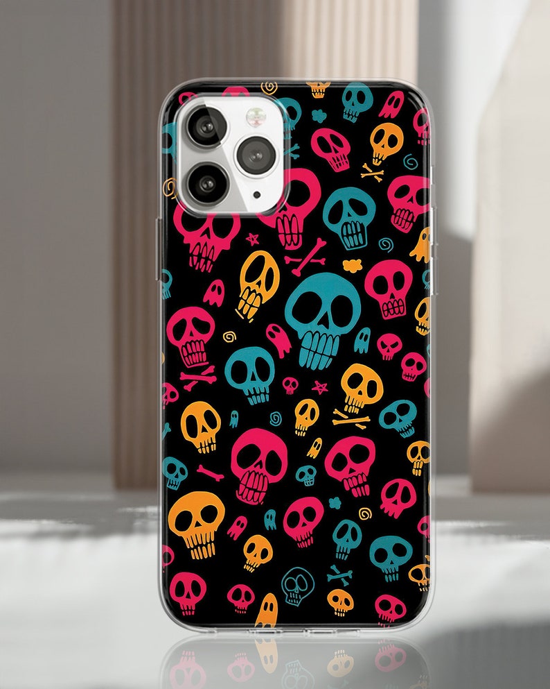 Puede incluir: Una funda de tel&eacute;fono negra con un patr&oacute;n repetido de calaveras de dibujos animados coloridas en rosa, amarillo, azul y naranja. La funda tiene un borde transparente y cubre la parte trasera del tel&eacute;fono.