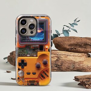 May include: Clear phone case with an orange retro game controller design. The case features a screen displaying a colorful space scene. The design includes a directional pad, buttons, and circuit board details, offering a nostalgic aesthetic.