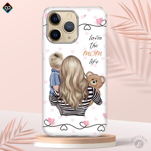 May include: A clear phone case with a pink and white background. The case features a cartoon illustration of a woman with blonde hair holding a teddy bear and a young boy. The text "lovin the mom life" is printed on the case.
