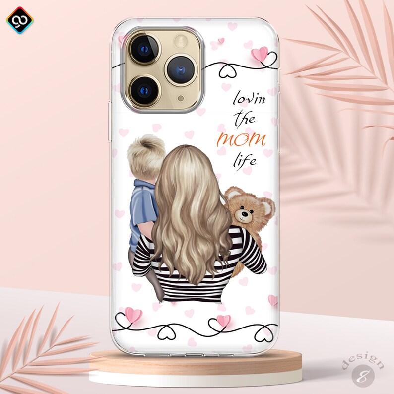 May include: A clear phone case with a pink and white background. The case features a cartoon illustration of a woman with blonde hair holding a teddy bear and a young boy. The text "lovin the mom life" is printed on the case.