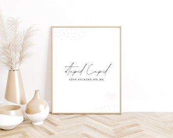 May include: A framed print with a white background and black text that reads "Stupid Cupid Stop Picking On Me". The print is surrounded by a light wood frame and is sitting on a wooden floor.